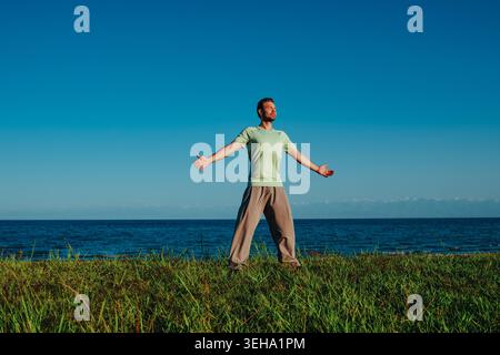 Mann in Sportbekleidung, der auf Gras steht, die Arme zur Sonne gestreckt, die Augen geschlossen Stockfoto