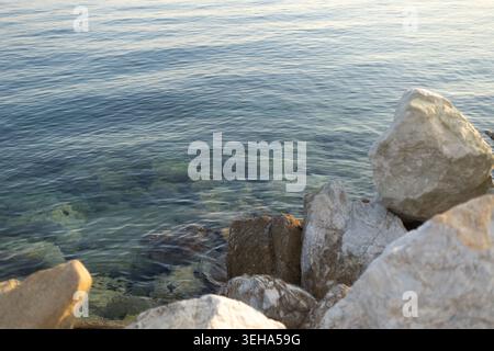 Helles klares Meerwasser, das sanft in der Nähe großer Felsen plätschert. Sichtbare Unterwassersteine im transparenten Ozean. Natürliche Küstenumgebung mit scharfem DE Stockfoto