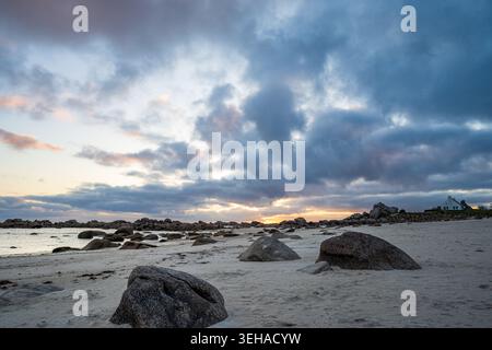 Ein malerischer Sonnenaufgang beleuchtet den Meneham Strand in der Bretagne. Große Granitblöcke sind auf weißem Sand verstreut, während im B ein typisches bretonisches Haus erscheint Stockfoto
