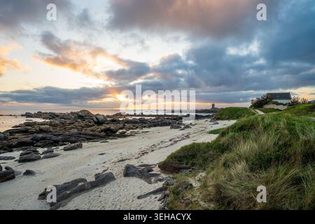 Ein spektakulärer Sonnenaufgang beleuchtet die zerklüftete Küste von Finistère. Warmes Licht filtert durch Wolken über dem weißen Sandstrand und charakteristischen Granitfelsen Stockfoto