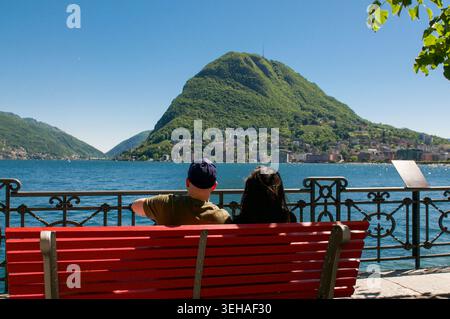Rückansicht eines jungen Paares, das auf einer klassischen roten Bank an der Seepromenade sitzt und den malerischen Panoramablick auf Mount San Salvatore und La genießt Stockfoto