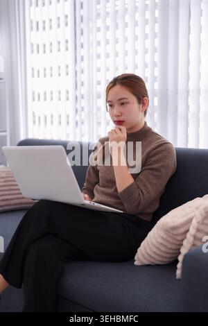 Junge asiatische Geschäftsfrau, die am Laptop arbeitet, während sie im modernen Büro auf dem Sofa sitzt und über die Geschäftsstrategie nachdenkt Stockfoto