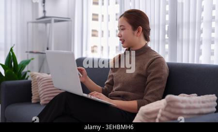 Junge asiatische Geschäftsfrau mit Laptop für Fernarbeit im Home Office, die auf einem modernen grauen Sofa in einer hellen Wohnung sitzt Stockfoto