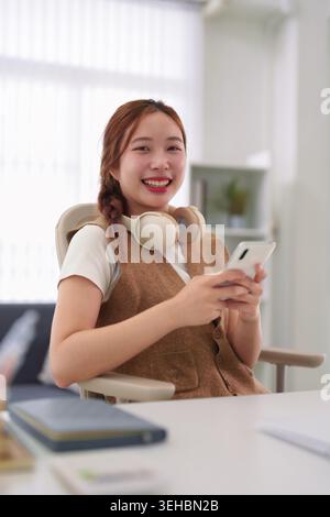 Junge asiatische Geschäftsfrau mit Smartphone, die online vom Schreibtisch im Heimbüro arbeitet, lächelnd fröhlich, fröhlich, professionell Stockfoto