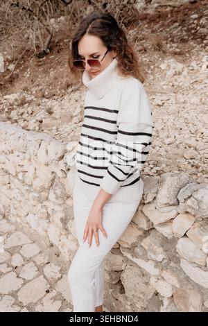 Der Lifestyle eines weiblichen Pullovers posiert an einem ruhigen Herbsttag in trendigen gestreiften Strickwaren und weißen Hosen vor einer Steinmauer in den Bergen Stockfoto