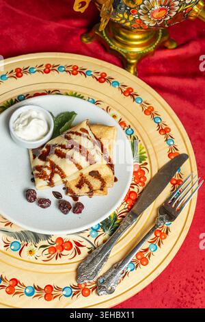 Goldene Crepes mit Schokoladennebel auf dekorativem Teller mit traditionellen Ornamenten, Sauerrahm und Vintage-Besteck Stockfoto