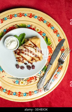 Gefaltete Blini mit Schokoladensauce, Sauerrahm und getrockneten Früchten auf traditionellem bemaltem Teller mit Vintage-Besteck. Perfekt für russische Kultur und h Stockfoto