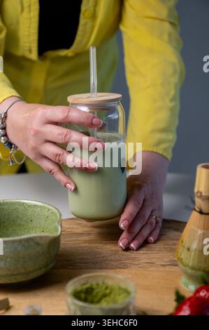 Frau in gelbem Mantel hält sanft Glas Matcha Latte Glas neben Keramikschale und Bambusquirl. Eiskaltes grünes Getränk mit Strohhalm in gemütlicher Küche Stockfoto