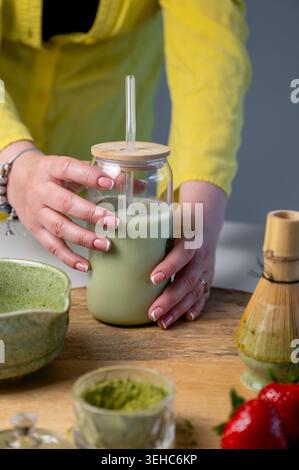 Kaukasische Hände gießen schaumigen Matcha Latte in ein Glas mit cremigem Schaum und Spritzen. Keramikschale und Bambusquirl auf Tablett mit grünem Pulver. Hausgemacht Stockfoto