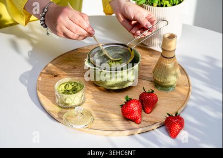 Lebendiges Matcha-Tee-Frühstück mit frischen Erdbeeren und Zubereitungswerkzeugen in hellem natürlichem Sonnenlicht. Hände-Sieben-Pulver mit energetischem Mor Stockfoto