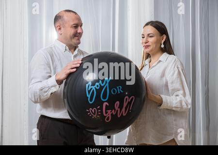 Weißes Paar Hält Geschlechterparty Ballon, Großer Schwarzer Ballon Liest Junge Oder Mädchen, Werdende Mutter Und Vater Lächeln In Lässigen Weißen Hemden, Stehend Stockfoto