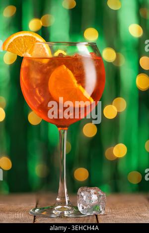 Aperol spritzt Cocktail in einem festlichen Glas auf rustikalem Holztisch mit funkelnden Bokeh-Lichtern in leuchtendem grünem Hintergrund Stockfoto