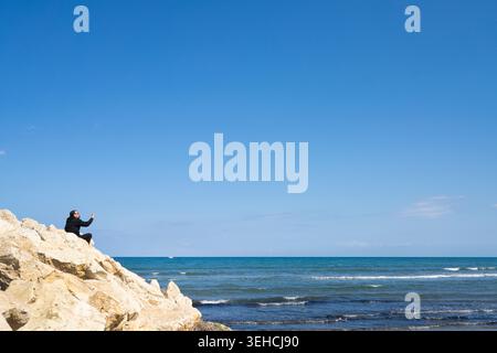 Larnaka, Zypern. Mai 2026. Ein Mann in einer schwarzen Jacke sitzt auf einem Felsvorsprung mit Blick auf das Meer und hält sein Handy hoch. Der Himmel ist ein klarer, heller bl Stockfoto