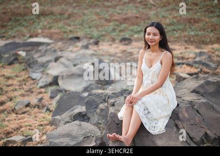 Frau in weißem Spitzenkleid, barfuß auf zerklüfteten Felsen sitzend, ruhiges Naturporträt mit weichen Erdtönen, friedlicher Sommer-Lifestyle, Entspannung im Freien Stockfoto