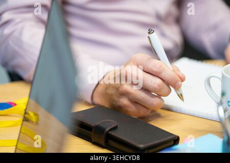 Ein Geschäftsmann schreibt auf einem Notizblock mit seinem Handy auf dem Schreibtisch neben ihm, ein Laptop im Vordergrund Stockfoto