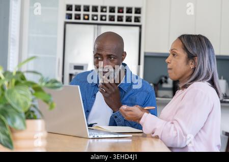 Seniorenpaar sitzt auf Kücheninsel und diskutiert Rechnungen auf silbernem Laptop mit offenem Notebook. Duo, Budgetierung, Planung, Zusammenarbeit, Homeoffice, modern, pflanzlich Stockfoto