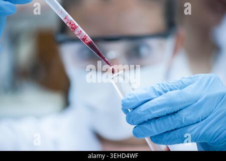 Ein Labortechniker aus dem Nahen Osten in Handschuhen, Maske und Schutzbrille pipettiert eine Blutprobe in ein Reagenzglas, während ein junger arabischer Arzt aufmerksam in einem klinischen Labor beobachtet, das sich auf Diagnostik und Forschung konzentriert. Stockfoto