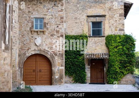 Vieste - Apulien - Italien - 08-29-2025: Verwitterte Steinfassade mit einem bogenförmigen Holztor, einer mit Efeu bedeckten Wand, einer kleinen Holztür unter einer gefliesten Markise und zwei Fenstern darüber. Stockfoto