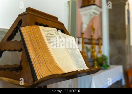 Sao Bras de Alportel, Portugal. Mai 2026. Die offene Bibel liegt auf einem geschnitzten hölzernen Rednerpult in einer Kirche mit Kerzen, Altardetails und Säulen Stockfoto