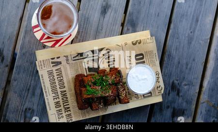 Blick von oben auf panierte frittierte Snacks, serviert mit weißer Dip-Sauce auf Holzbrett mit Zeitungspapier und ein Glas Bier auf einem rustikalen Tisch im Freien. Stockfoto