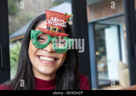 Frau lächelt, trägt grüne Glitzerbrille mit rotem Weihnachtsmütze und Creolen-Ohrring im Wohnzimmer Stockfoto