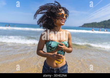 Junge Frau mit lockigen Haaren in bunten Sonnenbrillen und Bandeau-Top, die einen Ananasdrink an einem sonnigen Strand hält, lächelt, während die warme Brise und Wellen kreieren Stockfoto
