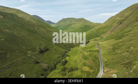 Ein Blick aus der Vogelperspektive fängt eine enge Asphaltstraße ein, die sich durch ein lebhaftes grünes Tal schlängelt, umgeben von sanften, grasbewachsenen Hängen und weit entfernten Gipfeln Stockfoto