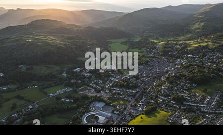 Ein ausladender Drohnenschuss fängt ein malerisches Dorf ein, das von üppigen, baumbedeckten Bergen umgeben ist, die in den warmen Glanz der untergehenden Sonne getaucht sind Stockfoto