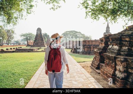 Weibliche Touristen mit rotem Rucksack und Strohhut, die auf einem Backsteinpfad spazieren, während sie die antiken Ruinen des Tempels Wat Ratchaburana in Ayutthaya erkunden Stockfoto