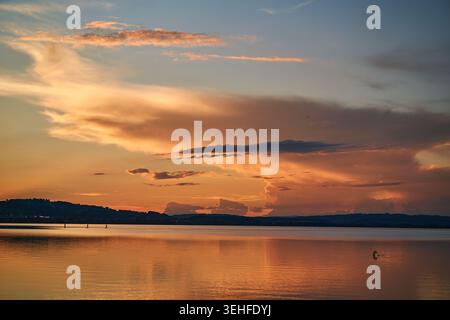 Warmer goldener Sonnenuntergang über einem ruhigen See, dramatische Wolkenlandschaft und spiegelartige Reflexe über ruhigem Wasser. Stockfoto