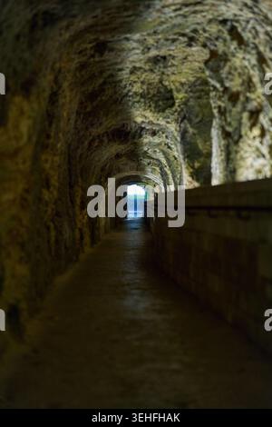 Langer, schmaler unterirdischer Tunnel mit zerklüfteten Steinmauern und einem Gehweg, der zu einem hellen Ausgang in der Ferne führt Stockfoto