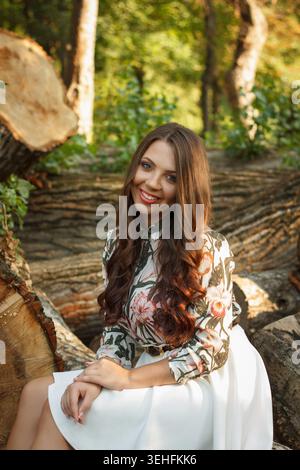 Eine junge Frau mit langen braunen Haaren sitzt auf einem Baumstamm in einem Wald. Sie trägt eine Bluse mit Blumenmuster und einen weißen Rock und lächelt in einer natürlichen Umgebung Stockfoto