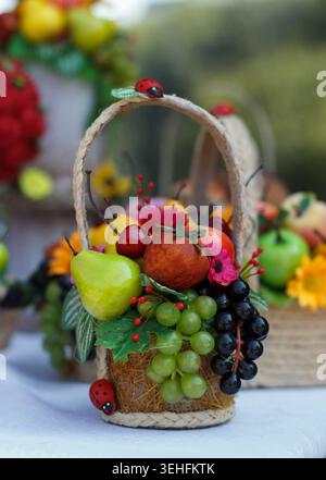 Ein dekorativer Korb gefüllt mit verschiedenen Früchten wie Trauben, Äpfeln, Birnen und Beeren. Der Korb ist mit grünen Blättern und Marienkäfern geschmückt Stockfoto