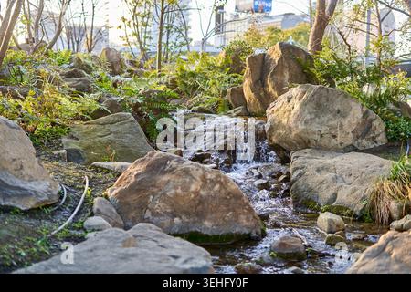 Eine ruhige Landschaft mit einem kleinen künstlichen Wasserfall und einem Bach, der durch große Naturfelsen in einer städtischen japanischen Gartengestaltung fließt. Der Stockfoto