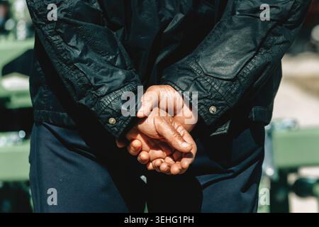 Ein älterer Mann steht mit den Händen hinter dem Rücken, trägt eine dunkle Jacke und eine marineblaue Hose. Die ruhige, kontemplative Pose lässt Geduld vermuten Stockfoto