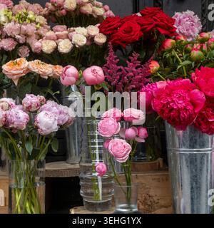 Eine lebendige Ausstellung von frischen Blumen, die in Glas- und Metallvasen an einem Marktstand angeordnet sind, mit rosa Pfingstrosen, roten Rosen, pfirsichfarbenen Sprayrosen, Stockfoto