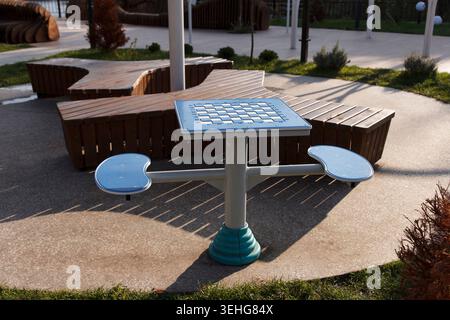 Ein moderner Schachtisch im Freien mit zwei Sitzplätzen in einem Park. Der Tisch ist mit einem Schachbrettmuster versehen. Die umliegenden Bänke bestehen aus Holz Stockfoto