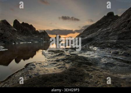Zerklüftete Felsen säumen eine ruhige Küste, die die warmen Farben des Sonnenuntergangs reflektieren Stockfoto