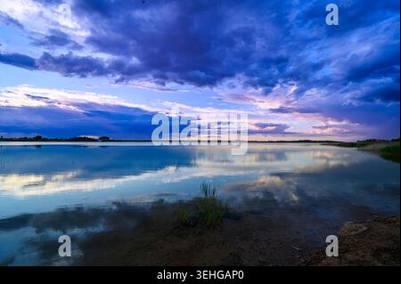 Dramatischer Himmel spiegelt sich perfekt auf ruhiger Seenoberfläche. Violette Wolken wirbeln über der fernen Horizontlinie. Sanft kräuselndes Wasser reflektiert die lebendigen Farben des Himmels. Stockfoto