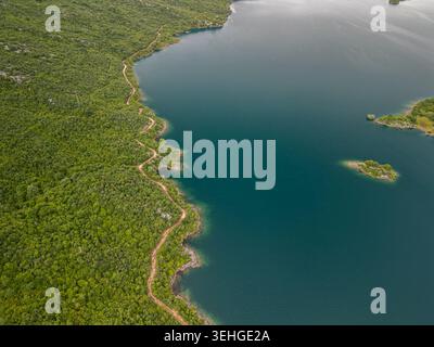 Slansko Lake, Montenegro - 05 29 2025: Abgewinkeltes Drohnenfoto, das einen ruhigen, türkisfarbenen See zeigt, der von dichtem Wald umgeben ist, eine gewundene Schotterstraße und zwei Inseln Stockfoto