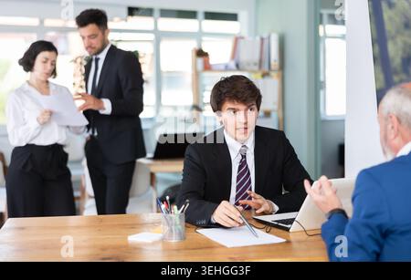 Erfahrener junger Bankmanager, der mit einem Reifen man-Kunden spricht und beim Unterzeichnen von Dokumenten im Büro hilft Stockfoto
