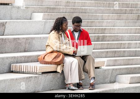 Ein nerdy-fokussierter schwarzer Mann erklärt der schwarzen Freundin, die draußen sitzt, Studien über Laptop. Stockfoto