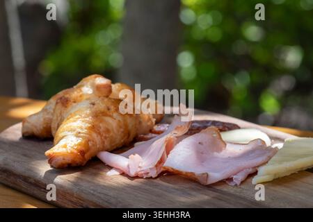 Rustikales Croissant mit Schinken und Käse auf hölzernem Servierbrett in der Brunch-Szene im Freien Stockfoto