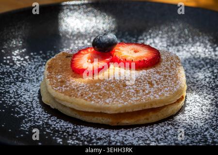 Flauschiger hausgemachter Pfannkuchen mit Erdbeerscheiben und Heidelbeere auf schwarzem Teller mit Puderzucker Stockfoto