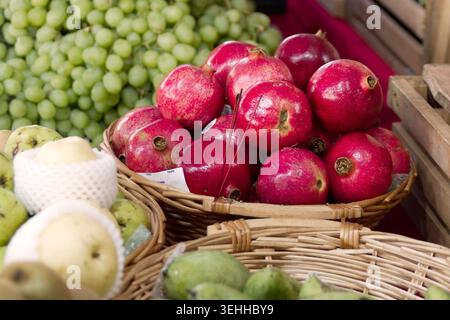 Ein Korbkorb gefüllt mit hellroten Granatäpfeln auf einem Markttisch neben grünen Trauben und gewickelten Birnen. Stockfoto