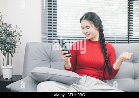 Junge asiatische Frau lächelt ihr Smartphone zu Hause auf dem Sofa an, überprüft Online-Shopping-App oder mobile Benachrichtigungen, digitalen Lebensstil der Verbraucher, E-Commerce br Stockfoto