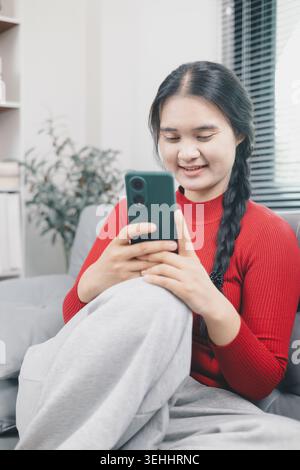 Junge asiatische Frau, die ihr Smartphone zum Online-Shopping zu Hause nutzt, mobile E-Commerce-App auf dem Sofa durchsucht, digitalen Lebensstil für Verbraucher, bequemer Kauf Stockfoto