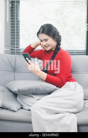 Junge asiatische Frau, die zu Hause ein Smartphone auf dem Sofa zum Online-Shopping nutzt, mobile E-Commerce-App durchsucht, digitalen Lebensstil für Verbraucher, bequemer Kauf Stockfoto