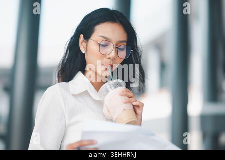 Nahaufnahme einer professionellen Frau mit Brille, die Eiskaffee trinkt, während sie Dokumente überprüft, ideal für Büroferien, Papierkram, geschäftliche Schwerpunkte und Stockfoto