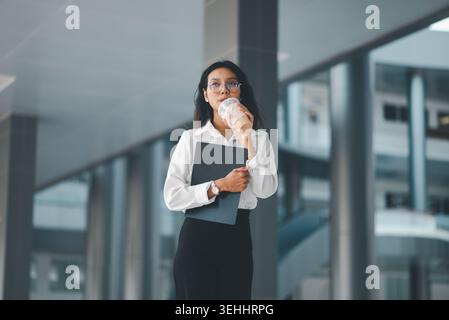 Professionelle Frau in Brille trinkt Eiskaffee, während sie ein Klemmbrett in einem modernen Bürogang hält, zeigt Büropause, ruhiges Selbstvertrauen, Bus Stockfoto